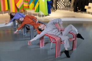 Kolme tanssijaa istuu kolmella perättäisellä raidallisella tuolilla taivutetussa  tanssiasennossa Antti + Vuokko -näyttelyssä.