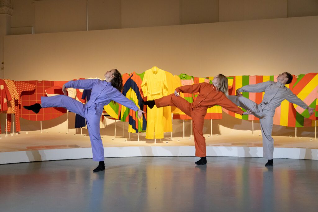Kolme tanssijaa seisovat rivissä ja kaikilla on kädet suorana sivuilla ja oikea jalka ylhäällä.